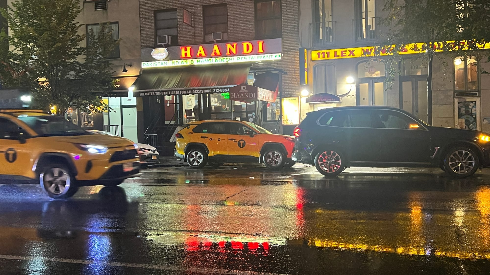 The red and white HAANDI sign lit up at night on a rainy Lexington Avenue, with yellow cabs and their reflections on the wet street.