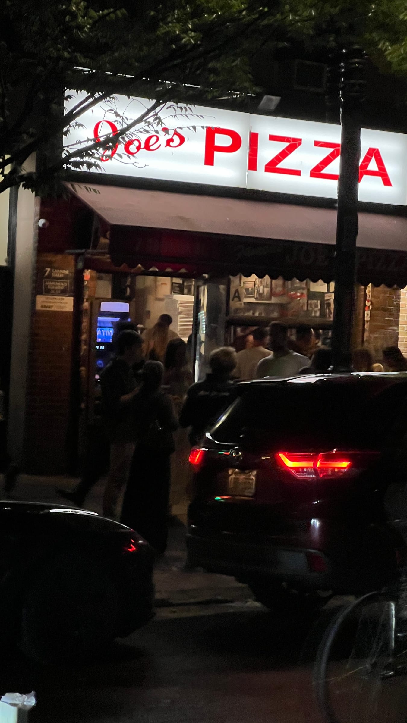 The red neon Joe’s Pizza sign glowing over a crowded counter at night, with people waiting on the sidewalk outside.