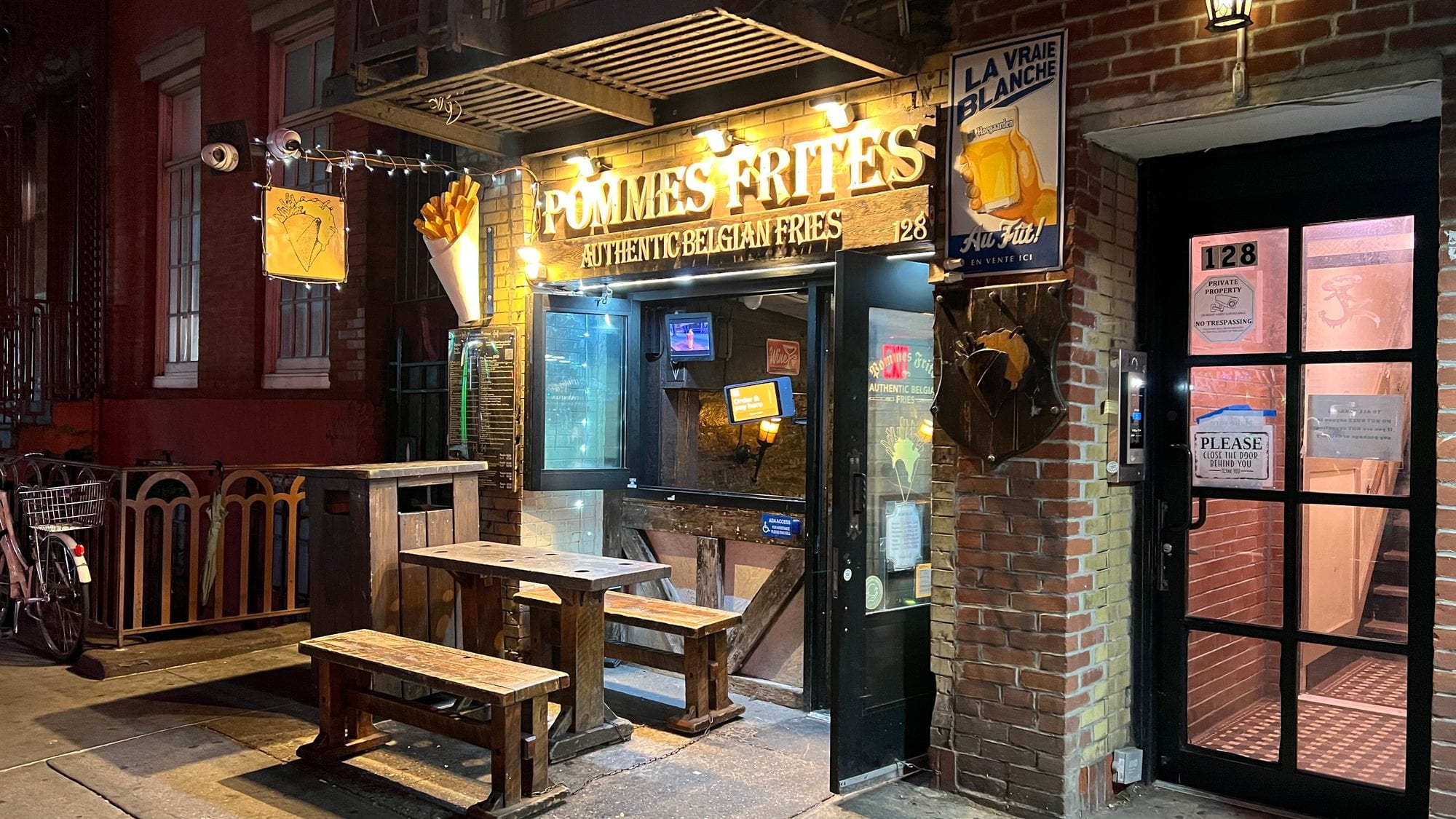The lit-up Pommes Frites storefront at night, with wooden benches out front and the Belgian fries sign over the door.