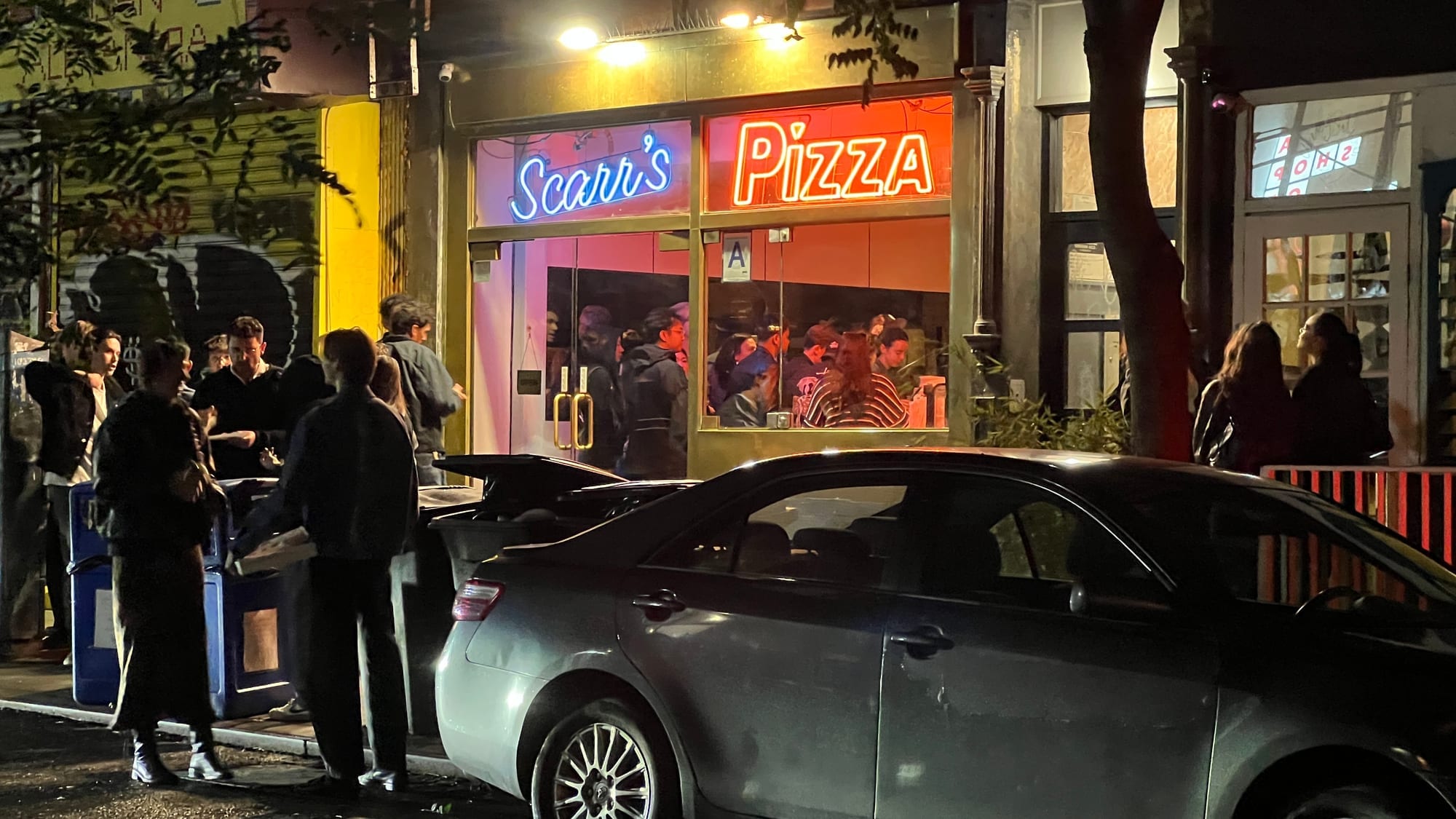 The neon Scarr’s Pizza sign in the window at night, with a crowd waiting on the sidewalk outside on Orchard Street.