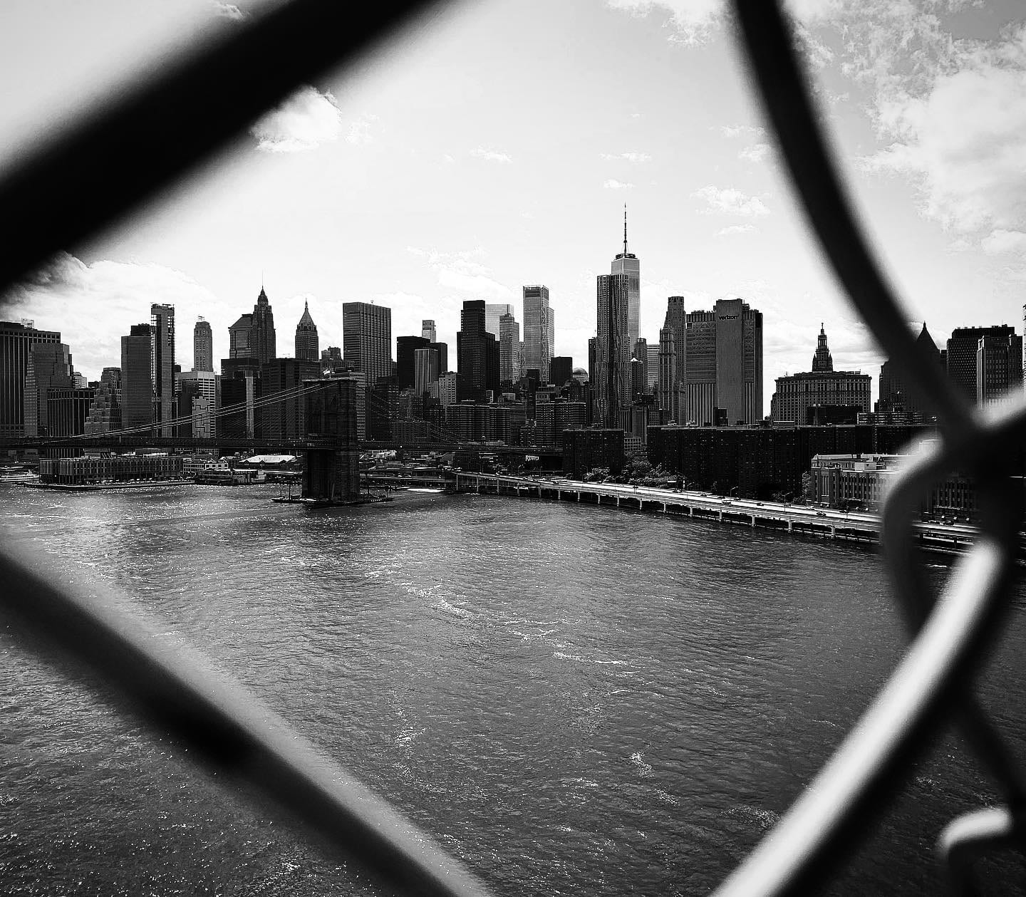 The Lower Manhattan skyline in black and white, shot through a hole in the Manhattan Bridge fence.