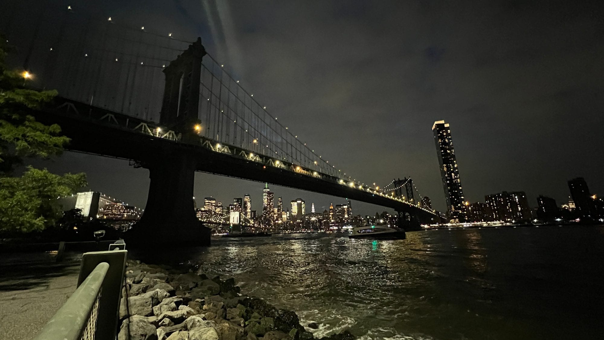 The Manhattan Bridge at night from the Brooklyn waterfront, with the lit Lower Manhattan skyline across the East River.