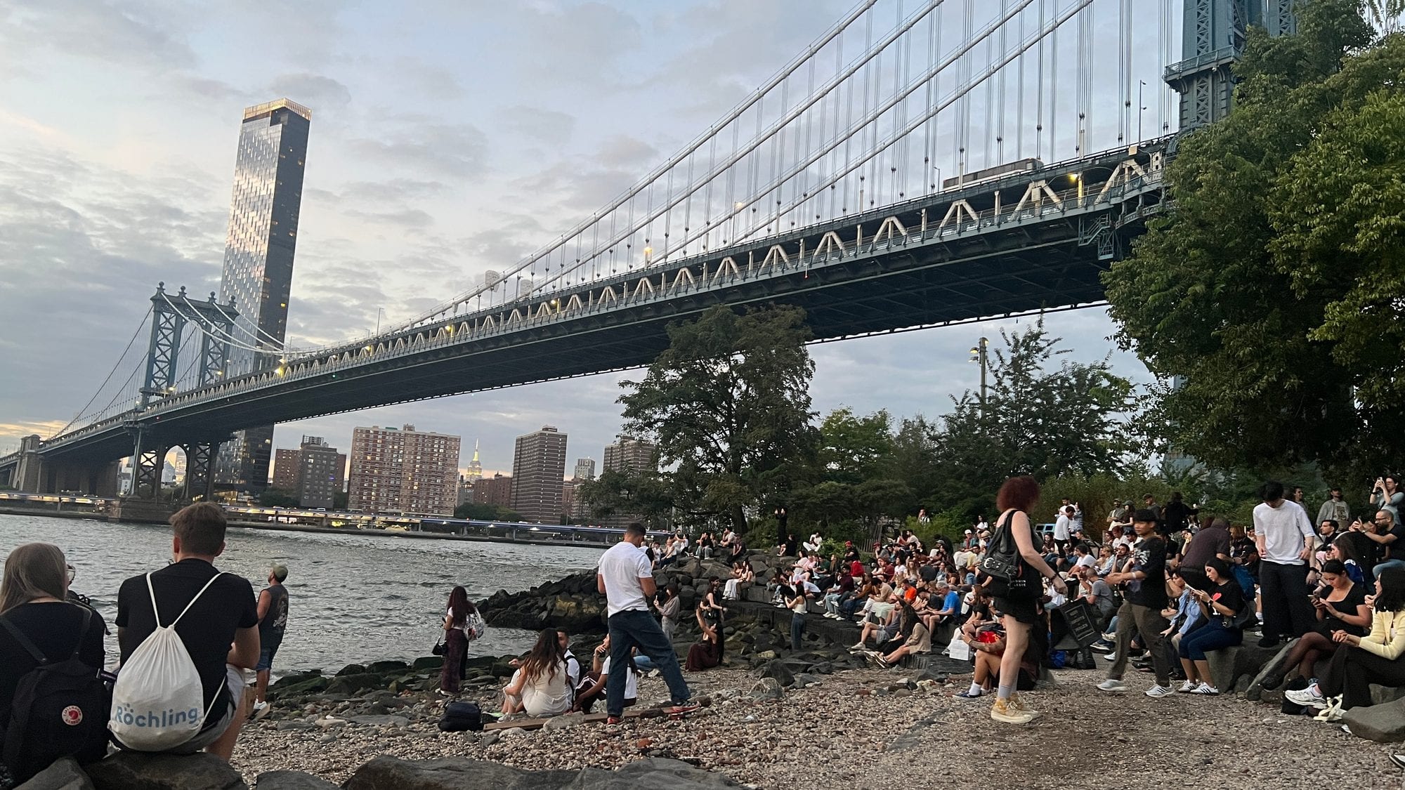 People sitting on the rocks under the Manhattan Bridge in DUMBO on a summer evening, one of the best free things to do in the city.