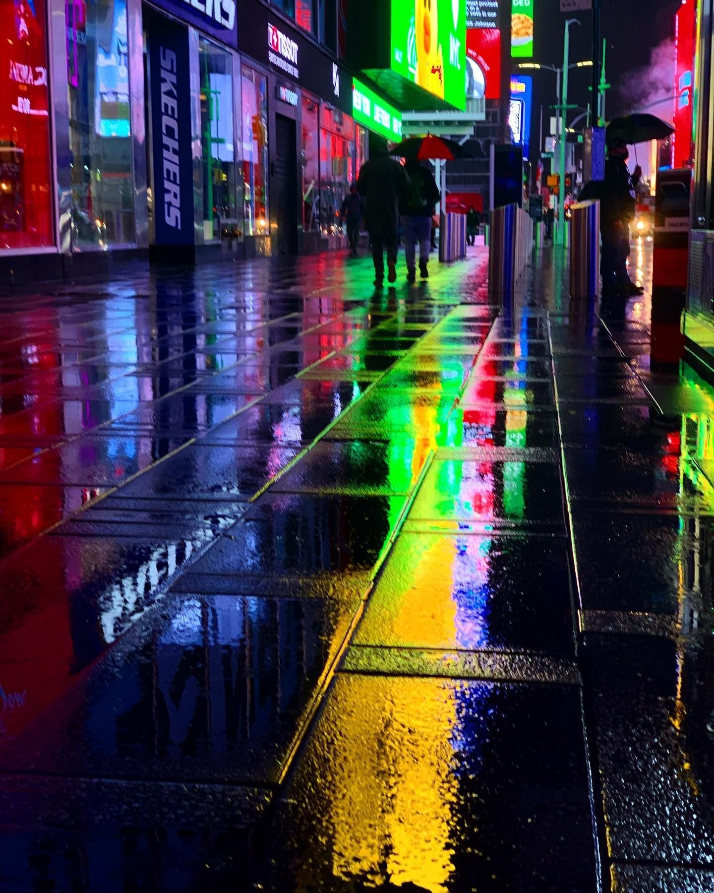 Times Square at night in the rain, the billboards reflected in bands of red, green and yellow down the wet sidewalk, with two people under an umbrella walking into it.