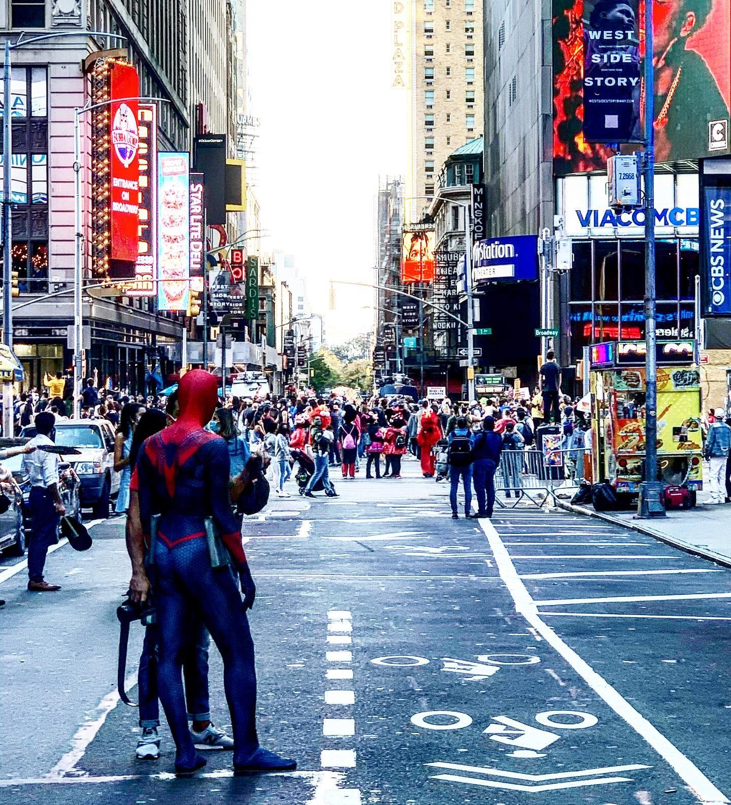 Times Square in daylight, crowds crossing Broadway with costumed characters working the sidewalk.