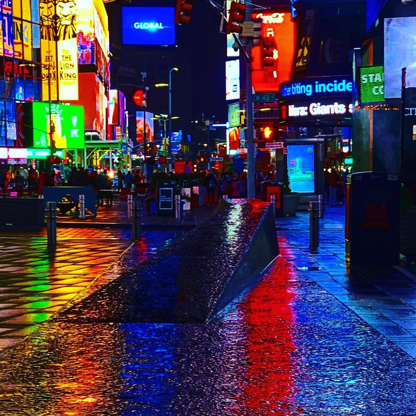 Times Square at night in the rain, the billboards reflected in red and blue across the wet pavement, with the crowd filling the block behind.