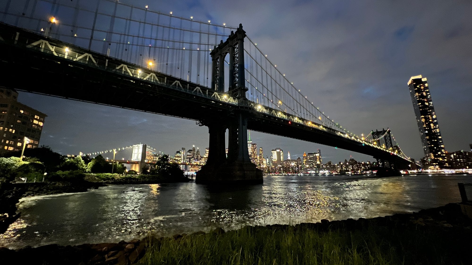 The Manhattan Bridge lit up at night from the DUMBO waterfront, with the Manhattan skyline glowing across the East River.