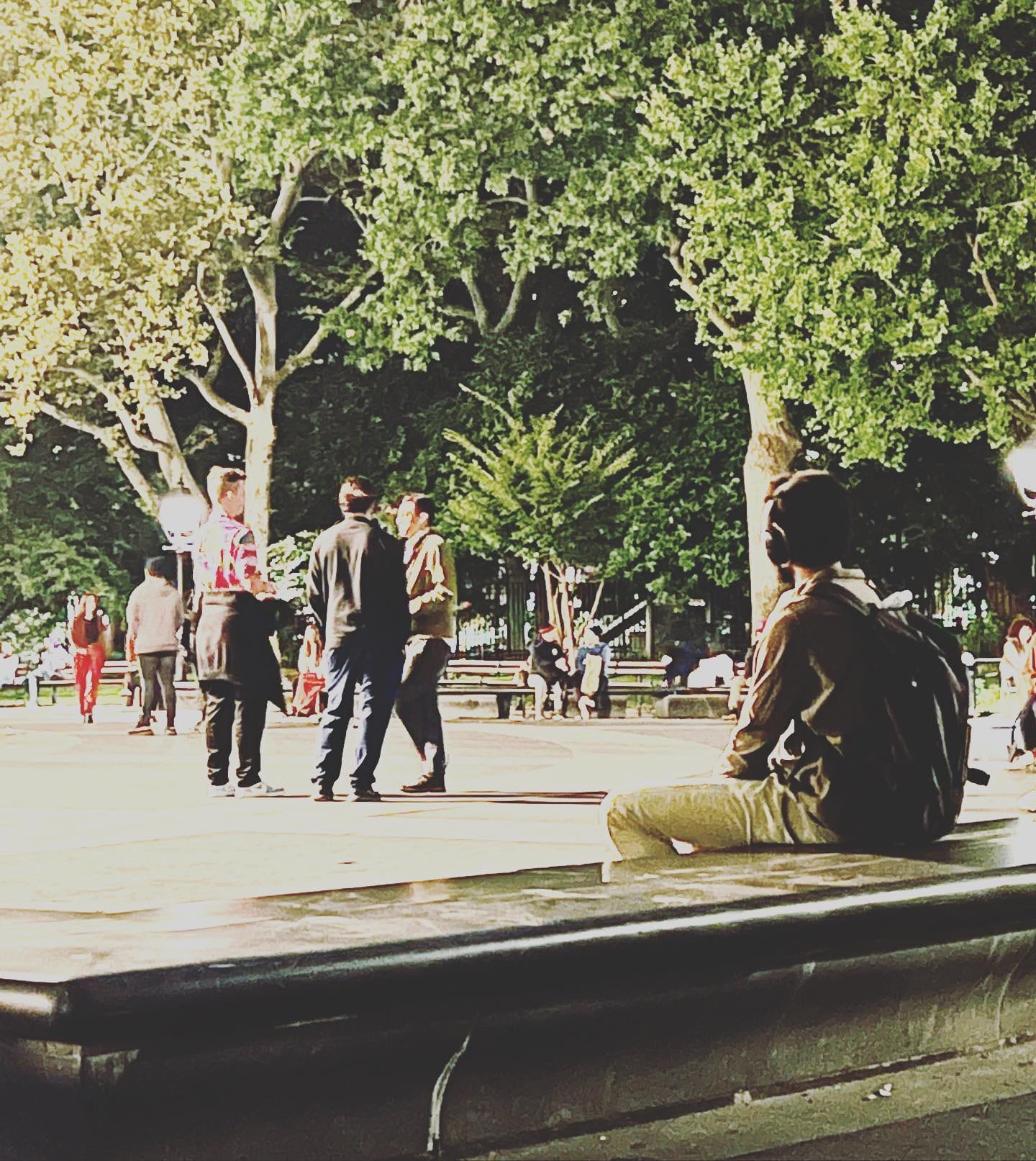 People standing and sitting on the fountain rim in Washington Square Park, under the plane trees at dusk.