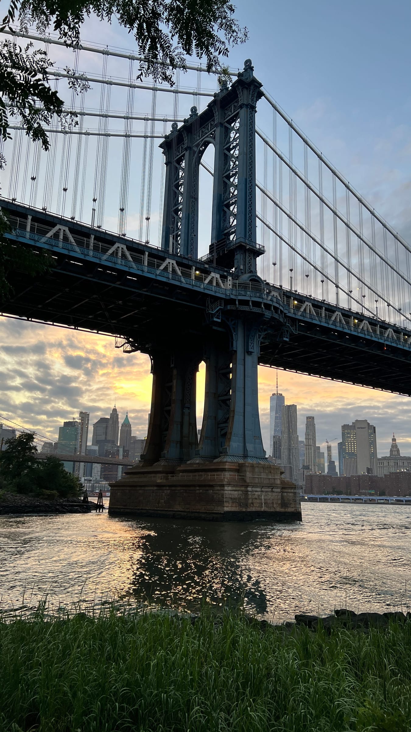 The Manhattan Bridge from the Brooklyn shore at sunset, its tower rising out of the East River with the Lower Manhattan skyline behind it.