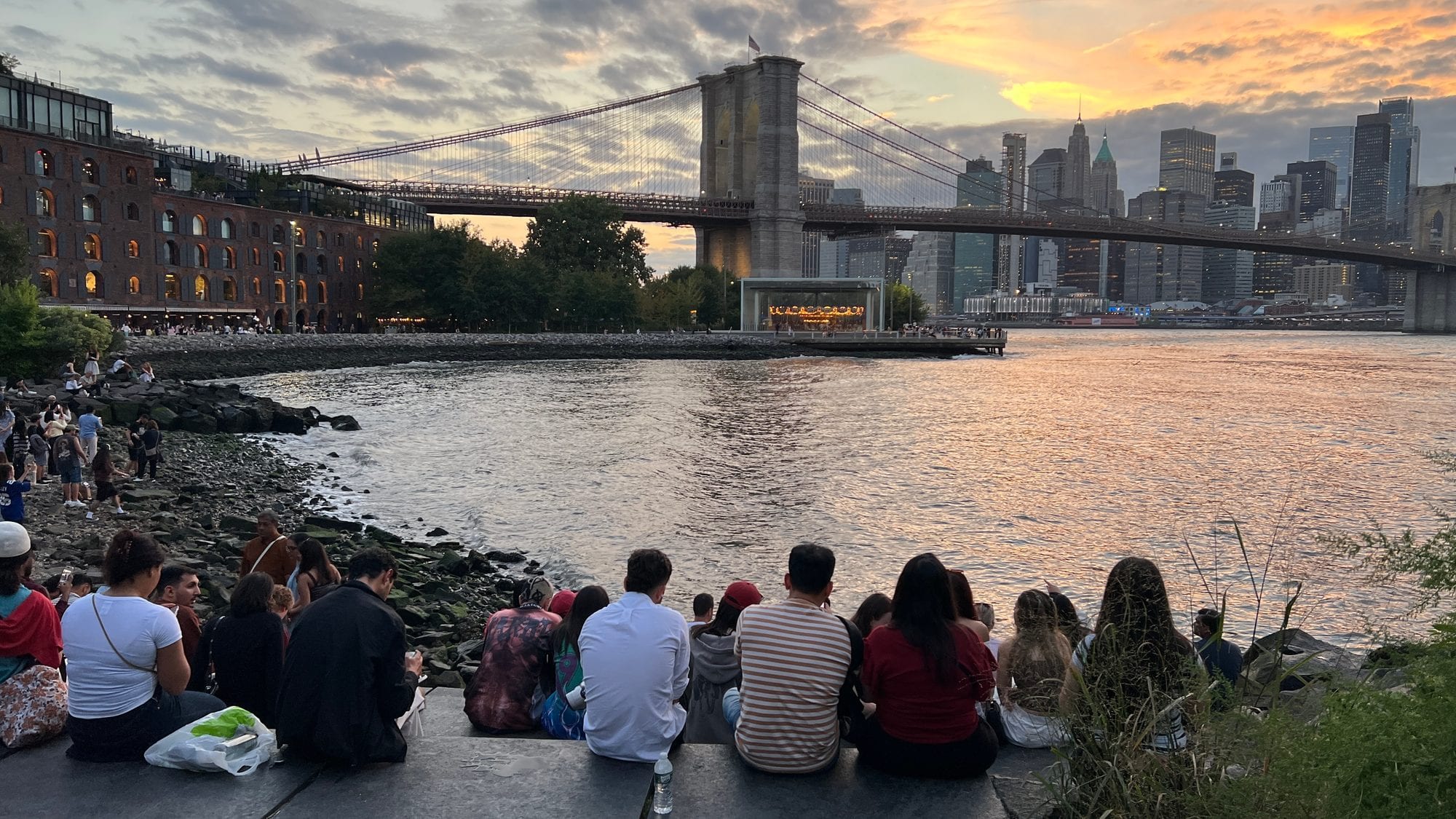 People sitting on the rocks at Pebble Beach in DUMBO at sunset, watching the Brooklyn Bridge and the Manhattan skyline across the water.