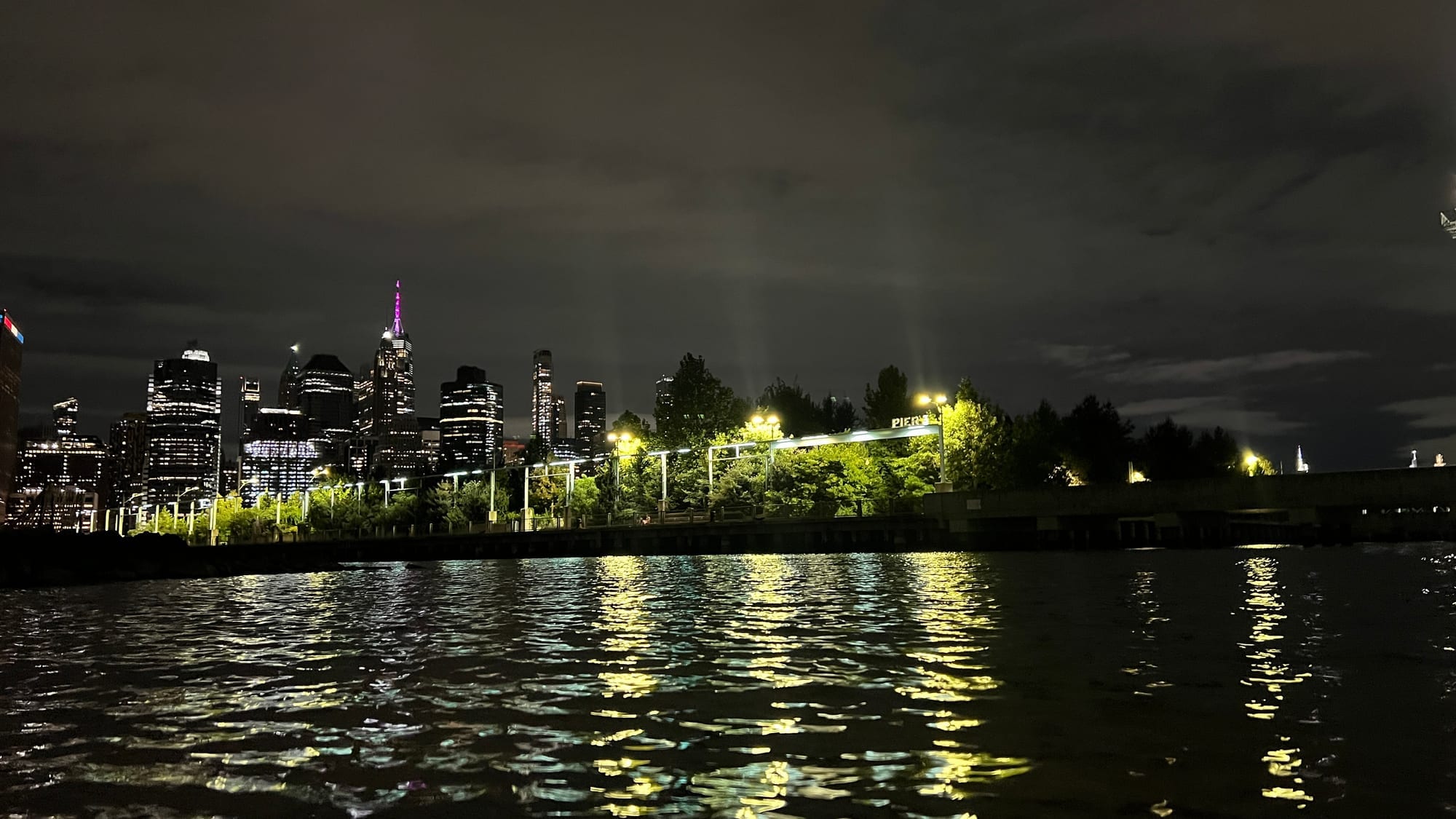 The lower Manhattan skyline at night, lit up across the dark East River, seen from Pier 3 in Brooklyn Bridge Park.