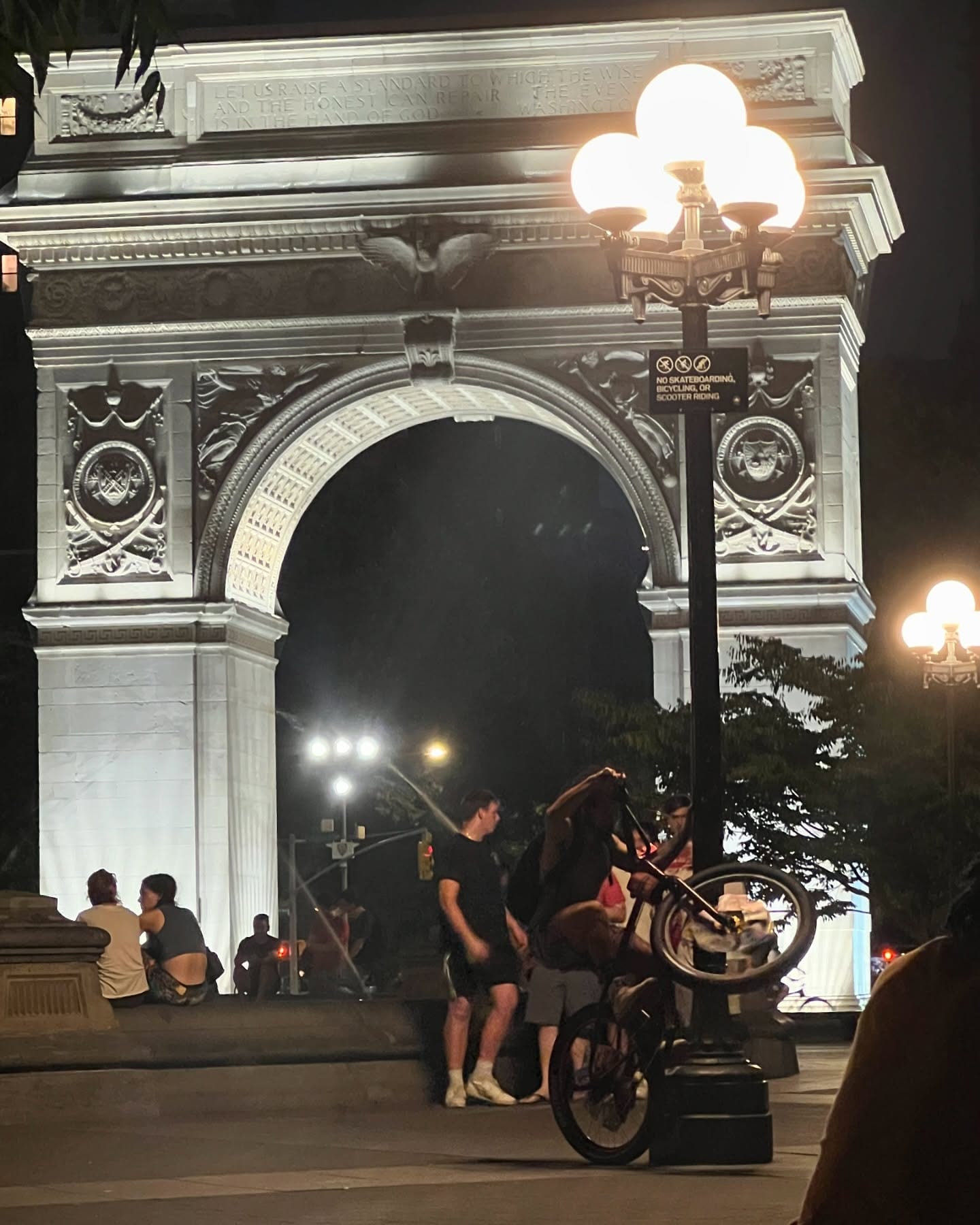 The Washington Square arch lit at night, with people gathered on the pavement beneath it.